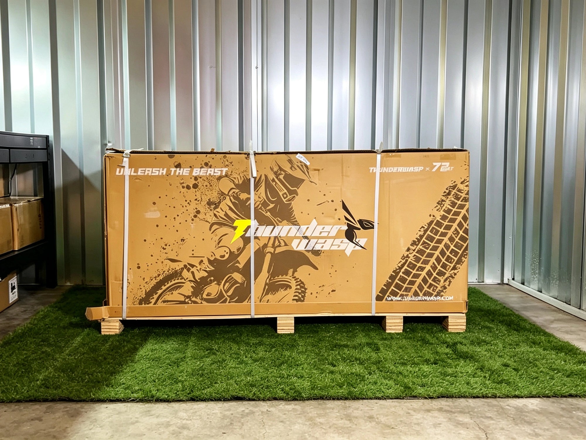 A large cardboard shipping box for Thunder Wasp, secured with white straps on a wooden pallet. The box features a brown graphic with a dirt bike and tire tread illustration.